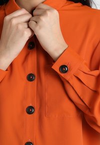 Orange skjorte med knaplukning foran, sorte knapper og krave, der foldes over. Har en brystlomme med en glat, tekstureret finish.