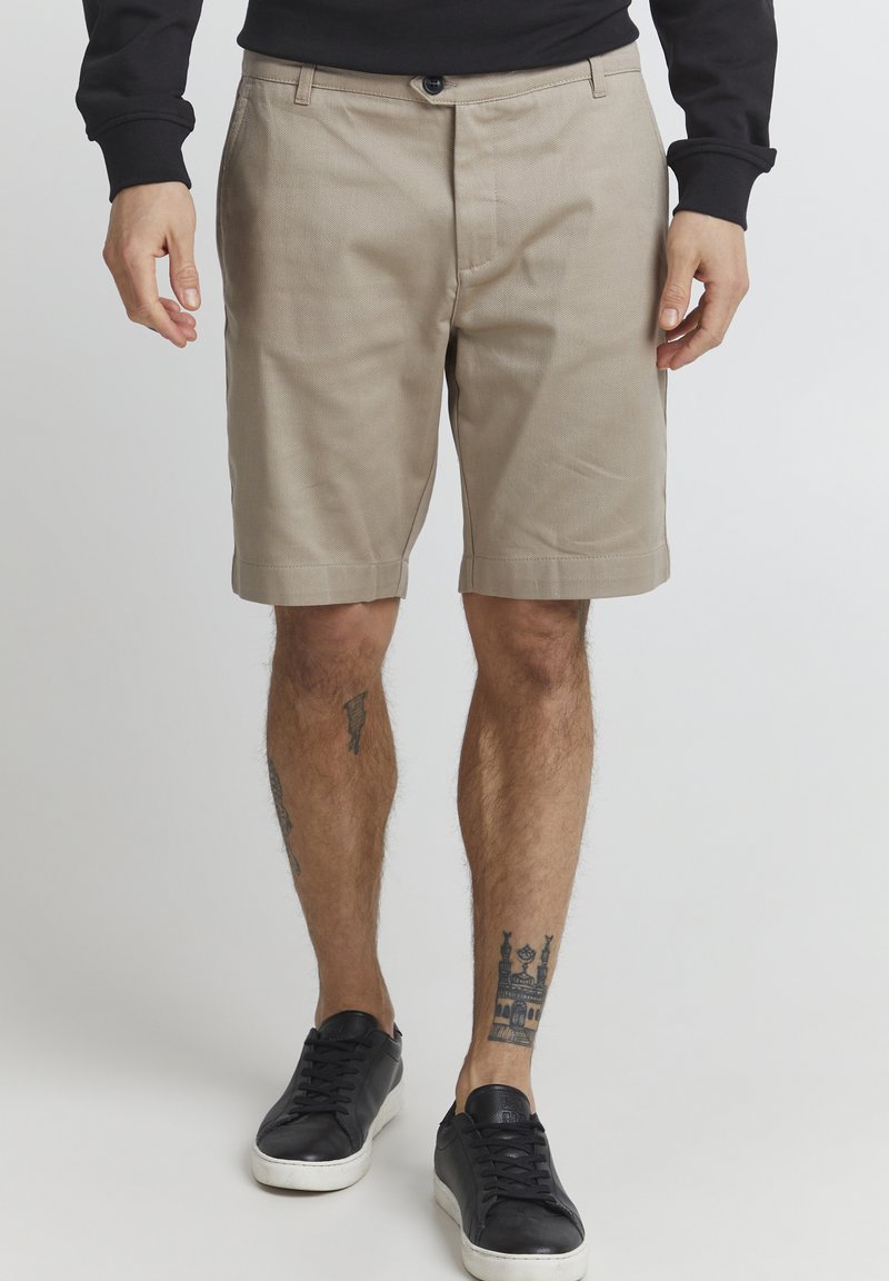 Tan shorts med flat frontdesign, knaplukning og sidelommer, lavet af et letvægtsstof. Sammen med sorte sneakers.