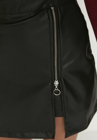 Jupe en simili cuir noire avec fermeture éclair sur le côté dotée de détails métalliques, et une fente ventée pour un design supplémentaire. Texture lisse sur l'ensemble.