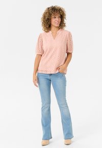 Light pink textured blouse with puff sleeves, featuring a V-neck and diamond patterns. Worn with light blue flared jeans and beige shoes.