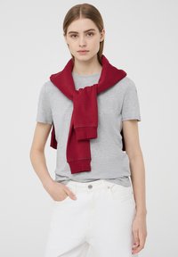 Maglietta grigia a maniche corte con un maglione rosso annodato attorno al collo. Modello che indossa jeans bianchi, mani nelle tasche, sfondo neutro.