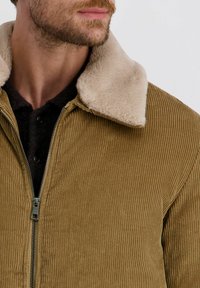 Bruine corduroyjas met een crème nepbont kraag, voorzien van een ritssluiting aan de voorkant en ribbelstructuur. Een zwarte geknoopte shirt is zichtbaar eronder.