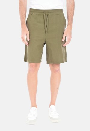 Uomo che indossa pantaloncini verde oliva con coulisse e scarpe da ginnastica bianche, in piedi contro uno sfondo chiaro uniforme.