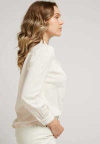 Blouse beige à manches longues en tissu texturé, col doux et détails froncés aux épaules, associée à un pantalon blanc.