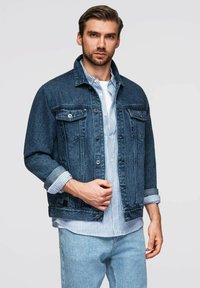 Giacca di jeans di colore blu scuro, con colletto classico, chiusura con bottoni, due tasche sul petto e maniche arrotolate, indossata sopra una camicia a righe.