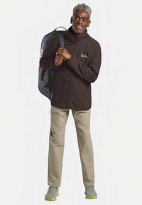 Braune Jacke mit Reißverschluss und Kragen, getragen über beigen Hosen. Die Figur trägt einen schwarzen Rucksack und graue Sportschuhe.