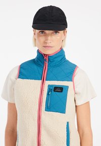Fleece vesta v modrej a krémovej farbe, so rúžovým zipsom, vreckom na hrudi a vysokým golierom. Čierna čiapka dopĺňa outfit.