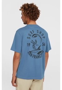 Light blue cotton t-shirt featuring a large black graphic of a surfer and text "SURF SHOP SAN FRANCISCO 1956" on the back. Short sleeves.