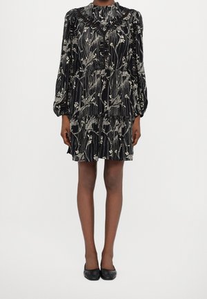 Kvinde iført en sort blomsterkjole med lange ærmer, flæsedetaljer og sorte slip-on sko mod en ensfarvet hvid baggrund.