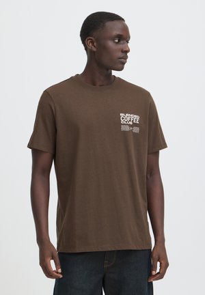Giovane uomo che indossa una maglietta marrone con il testo "Blended Coffee Club" sul petto, in piedi e guarda verso destra su uno sfondo semplice.