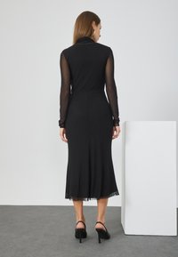 Μαύρο φόρεμα με μακριά διαφανή μανίκια, εφαρμοστό κορμάκι και φουσκωτό φούστα. Διαθέτει λεία υφή και διακριτική λεπτομέρεια σε στιλ στρωμάτων στη φούστα.