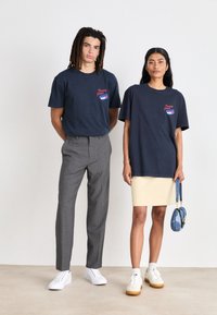 Camisetas de algodón marinas con logotipo rojo en el frente, combinadas con pantalones grises y una falda beige. Se presentan zapatillas y un pequeño bolso azul.