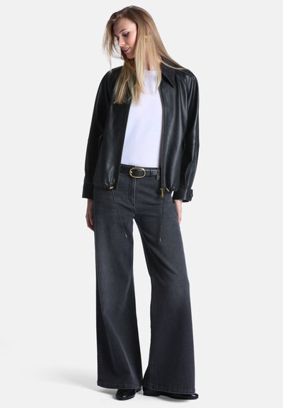 Jeune femme portant une veste en cuir noire, une chemise blanche, un jean noir à jambes larges avec ceinture, et des chaussures noires, debout et regardant vers le bas.