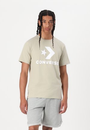 Světle khaki bavlněné tričko s velkým bílým logem Converse a designem hvězdy. V kombinaci se šedými šortkami s bílým lemem na okraji.