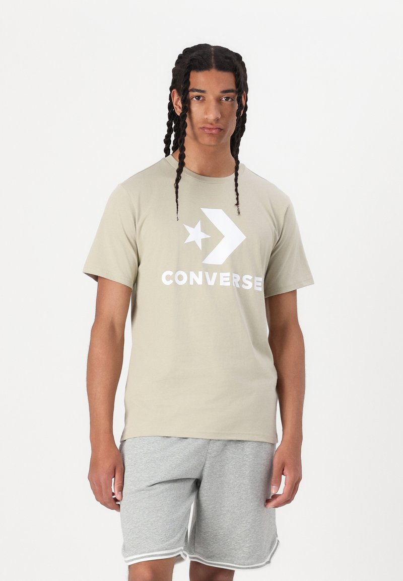 Gaišs haki krāsas kokvilnas t-krekls ar lielu baltu Converse zīmolu un zvaigznes dizainu. Apvienots ar pelēkiem šortiem ar baltu apmalīti ap apakšu.