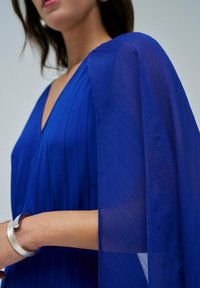 Vestido azul con pliegues y mangas transparentes, que presenta un diseño de escote en V. Una pulsera de plata adorna la muñeca, resaltando los detalles del conjunto.