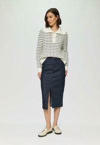 Gestreifter cremefarbener und marineblauer Pullover mit Kragen und Knöpfen, kombiniert mit einem dunkelblauen Jeansrock mit einem Schlitz vorne. Weiße Loafers runden das Outfit ab.