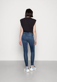 Svart ärmlös topp, figurnära mörkblå skinny jeans och vita sneakers med blå detaljer. Modellen vänder ryggen mot kameran och visar bakre detaljer.