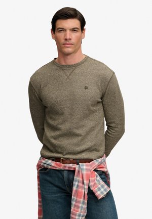 Homme aux cheveux foncés portant un pull marron et un jean bleu avec une chemise rose à carreaux nouée autour de la taille, sur un fond uni.