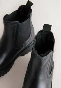 Dos botas de tobillo negras con una textura de cuero suave y paneles de tela, que cuentan con un forro acogedor y tiradores para facilitar su uso, colocadas sobre un fondo claro.