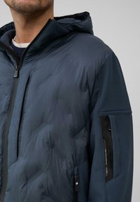 Kurtka puchowa w ciemnym granacie, z pikowanym przodem, fakturowanymi rękawami, kapturem, przednim zamkiem błyskawicznym i boczną kieszenią z subtelnym detalem logo.