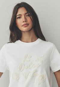 T-shirt en coton blanc avec une broderie en perles en relief portant le texte "La Dolce Vita", un étoile de mer et un motif de coquillage. Encolure classique.