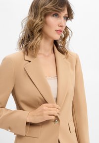 Femme au cheveux ondulés châtain clair portant un blazer beige sur un top crème bordé de dentelle, en train de fermer le bouton unique du blazer.