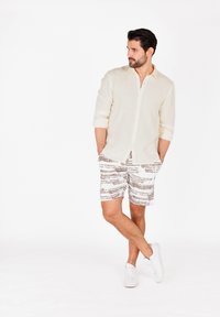 Camisa beige claro de botón con una tela texturizada, combinada con pantalones cortos blancos con rayas horizontales marrones. Llevada con zapatillas blancas.