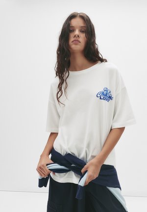 T-shirt blanc à manches courtes avec un design graphique bleu, associé à un pull bleu et bleu clair noué à la taille, porté avec un pantalon sombre.
