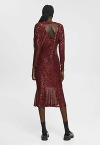 Lång röd paljettklänning med låg rygg, figurnära midja, utsvängd nederkant och långa ärmar. Bärs med svarta ankelsockor och skor.