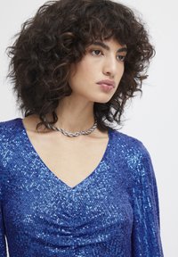 Blå strålande topp med V-ringning och långa ärmar, med samlade detaljer framtill och en slät textur. Silverhalsband i accent.