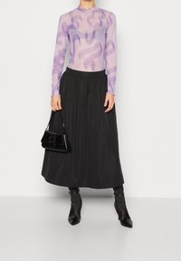 Langärmliges lila Mesh-Oberteil mit abstrakten Mustern, kombiniert mit einem schwarzen, fließenden Rock mit elastischem Bund und einer glänzenden schwarzen Handtasche.