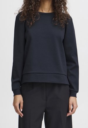 Femme aux cheveux bouclés portant un sweat-shirt noir uni à manches longues et un pantalon noir, debout devant un fond clair uni.