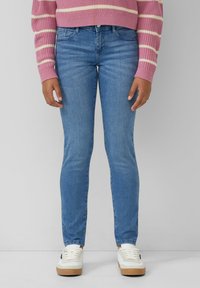 Tanke modre jeans hlače s rahlo elastičnostjo, ki imajo pet žepov in srednje visok pas, v kombinaciji z belimi supergami in roza črtastim puloverjem.