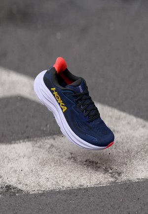 Chaussure de sport bleue avec une tige en mesh, une semelle intermédiaire blanche, un onglet de talon rouge et un logo jaune. Dotée de lacets noirs et de détails de surface texturés.