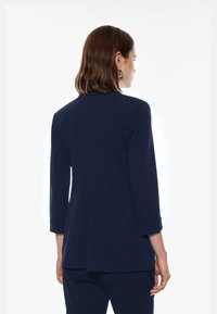 Blazer blu navy con maniche a tre quarti, vestibilità sartoriale e dettaglio con spacco sulla schiena, realizzato in un tessuto morbido e elastico.