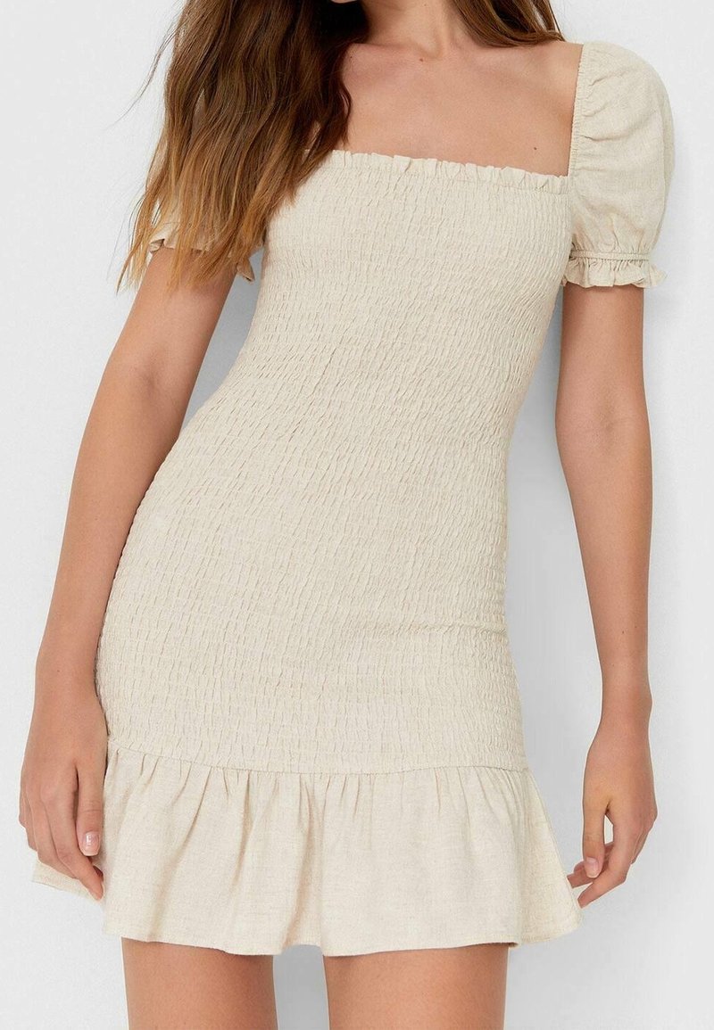 Robe beige ajustée avec un décolleté carré et des manches bouffantes. Tissu texturé avec un ourlet à volants pour plus de détails. Matière lisse et légère.