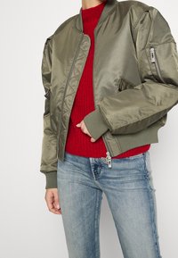 Chaqueta bomber verde oliva con un acabado satinado, puños acanalados y bolsillos laterales con cremallera, llevada sobre un suéter de punto rojo y jeans claros.