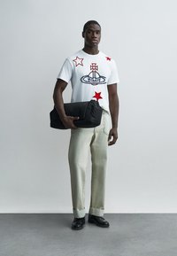 Mann som holder en svart sammenrullet bag, iført hvit t-skjorte med røde stjerner og grafikk, lyse bukser og svarte sko, står på en grå gulvflate.