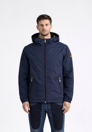 Uomo che indossa una giacca con cappuccio blu navy con cerniera frontale e due tasche con cerniera, abbinata a jeans bicolore blu, in piedi contro uno sfondo bianco.