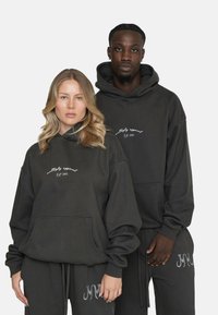 Dunkelgraue Hoodies aus weichem Stoff mit Kängurutaschen. Die Vorderseite ist mit einem weißen gestickten Logo versehen. Das Modell kombiniert mit passenden Jogginghosen.