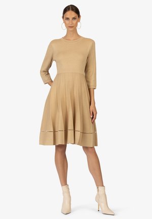 Robe tricotée jusqu'aux genoux en beige clair avec des manches trois-quarts, jupe plissée et découpes décoratives près de l'ourlet. Associée à des bottines beige.