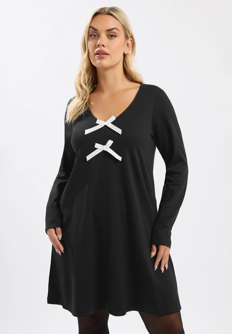 Schwarzes langärmliges Kleid mit V-Ausschnitt, das zwei weiße Schleifenakzente auf der Brust hat. Weicher Stoff, lockerer Schnitt, knielanges Design.