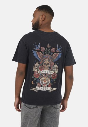 Uomo che indossa una maglietta nera con una grafica colorata sulla schiena che raffigura teschi, rose, una tigre ruggente, uccelli e la scritta "Brave Soul MCMLXXII".