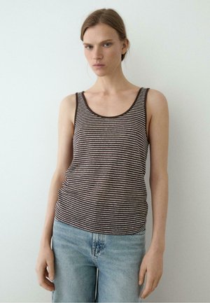 Młoda kobieta ubrana w brązowy i biały paskowany top na ramiączkach oraz jasnoniebieskie jeansy, stojąca na jednolitym jasnym tle.