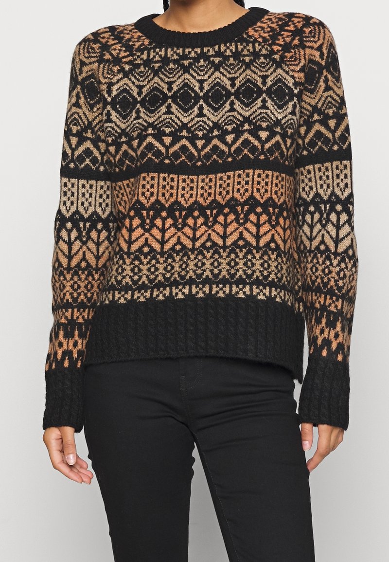 Femme portant un pull en maille noire avec des motifs géométriques et floraux marron et beige, assorti d'un pantalon noir, sur un fond neutre.
