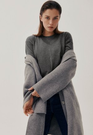 Ung kvinde med glat hår bagud iført en grå sweater og en oversized grå frakke, som justerer ærmet på frakken over mørke jeans.