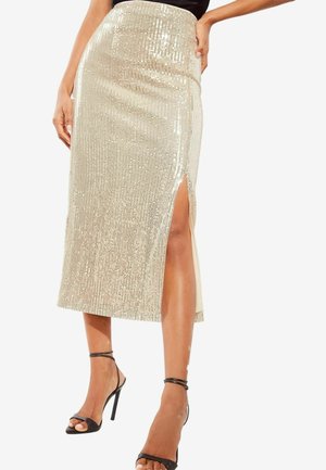 Femme portant une jupe midi en sequins dorés brillants avec une fente latérale et des sandales à talons noirs, debout avec une main sur la hanche.