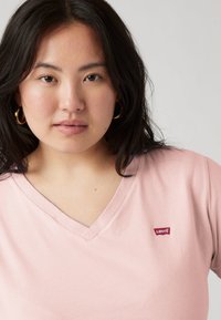 Ανοιχτό ροζ κοντομάνικη μπλούζα με λαιμόκοψη V από απαλό ύφασμα, με μικρό κόκκινο λογότυπο Levi's στο μπροστινό μέρος. Το ένδυμα έχει κοντά μανίκια και άνετη εφαρμογή.