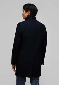 Cappotto in misto lana blu navy con colletto alto e cuciture visibili. Presenta un dettaglio con logo sul retro ed è tagliato dritto.
